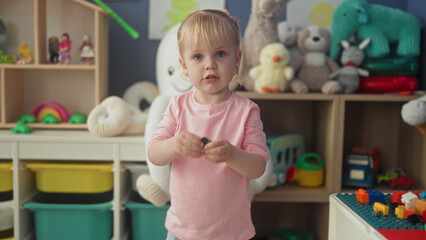 Fototapeta premium Little blonde girl holds a small toy block with both hands, hands extended toward camera in a play area inside a building; childhood curiosity.