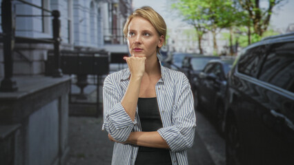 Fototapeta premium Woman with hand on chin and crossed arms on a street with parked cars and building facades; concern doubt decision.