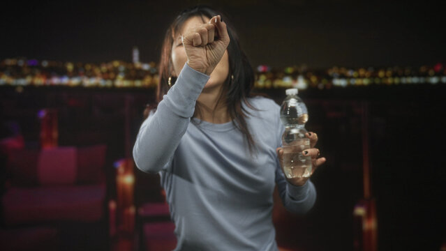 Woman holds water bottle up with hand while winking in studio against lit city skyline and lounge seating; playful confidence.