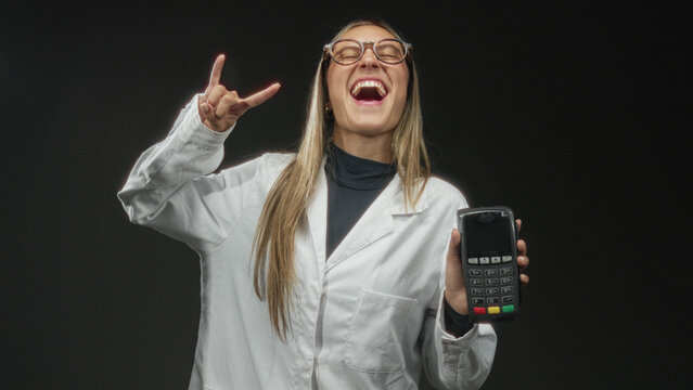 Woman wearing lab coat and glasses holding a payment terminal card reader and making rock horns gesture in studio; joyful confidence.