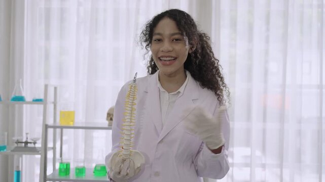 Happy professional woman medical anatomist wearing white lab coat uniform standing holding an anatomical model of the human spine with presentation and explanation carefully detailed in lab at clinic.