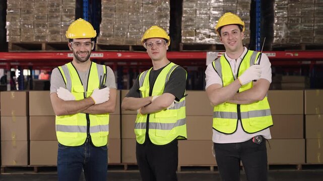 A group of Happy young Caucasian male workers in vest uniforms wearing yellow helmets is standing smiling and arms crossed while looking at the camera in a logistics warehouse distribution industry. 