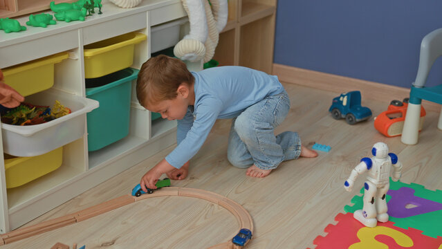 Boy kneeling pushing a toy car beside a blue white robot on foam puzzle mat near train track and storage shelf in building; playful curiosity.