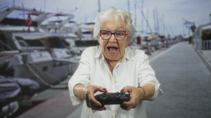 Senior woman holding gamepad with both hands pointed forward on a marina pier, smiling and reacting as she plays  playful joy. © Krakenimages.com