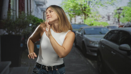 Fototapeta premium Woman dancing with hands raised and mouth open beside parked cars on a tree lined urban street wearing white tank top and jeans; joyful freedom.