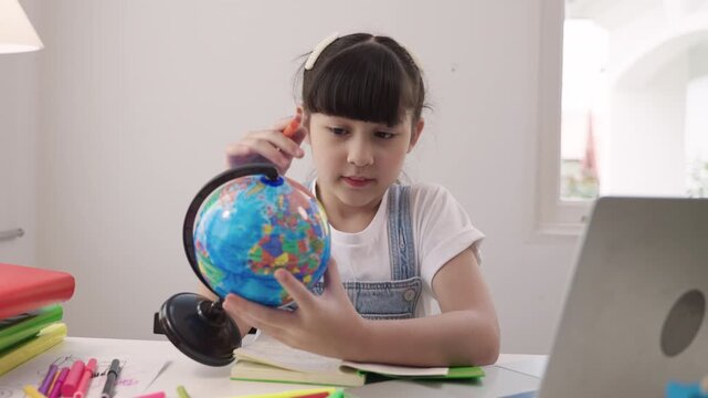 Cute Asian schoolgirl sitting at a desk using laptop computer for studying online learning via video calling and a remote teacher while having a geography lesson with a globe model in homeschool room.