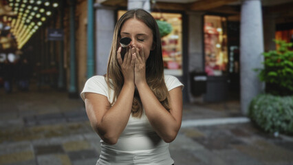 Fototapeta premium Young woman applying makeup brush to cheek, hands covering mouth gesture, brunette in white tee amid lit shopping street; surprise beauty vulnerability.