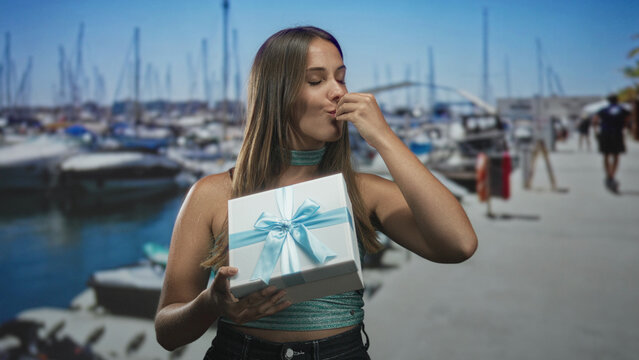 Young brunette woman holding white gift box with outstretched hand on street pier by moored boats and boardwalk; surprise celebration.
