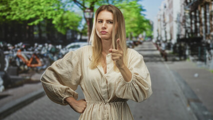 Fototapeta premium Blonde woman in linen dress standing with hands on hips and one finger raised on a street with bicycles and buildings behind her; determination.