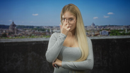 Obraz premium Woman touching glasses with finger at nose bridge on building terrace overlooking city; thoughtful contemplation.