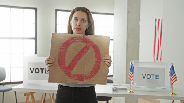 Woman young caucasian holding cardboard sign with red prohibition circle and points finger to sign in a voting building with ballot box and usa flags visible; defiance distrust.
