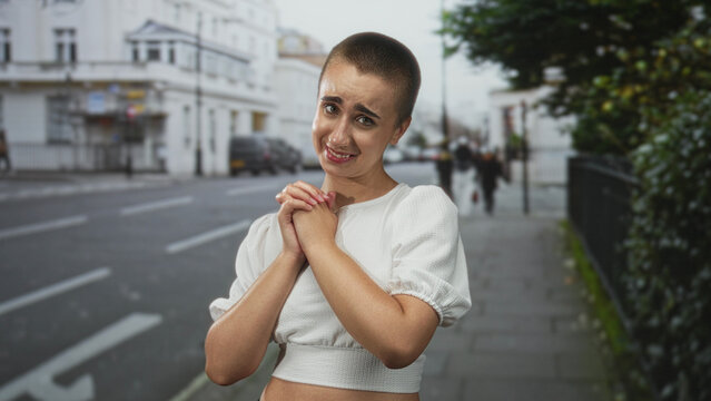 Woman with short buzzcut clasping hands to chest on street by row houses, white crop top and pleading tearful expression; vulnerability hope.