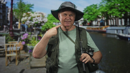 Fototapeta premium Man fisherman points finger to cheek while holding fishing rod and binoculars on street by a canal, wearing hat and vest; adventure.