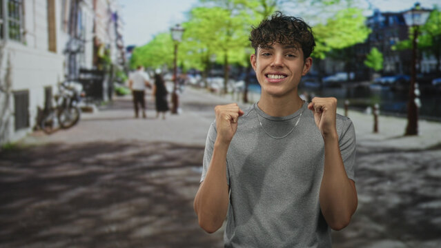Teenage man pointing fingers with bare hands toward camera, smiling and gesturing in a sunny street by a canal with bicycles and lamp posts; playful confidence.