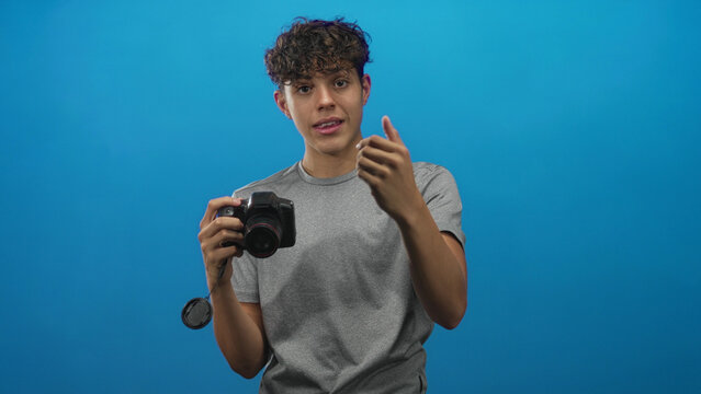 Teenage man holding dslr camera, pointing hand with fingers splayed while wearing gray tshirt in studio; curiosity study.