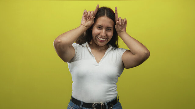 Woman smiling and making horn gesture with both hands near head in yellow studio wearing white top and jeans, casual expression; playful.