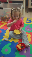 Naklejka premium Girl holding yellow spoon and plastic plate, stirs and lifts spoon to mouth while seated on number foam mat inside building; playful curiosity.