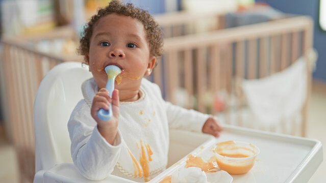 Little girl eating puree, holds spoon to mouth with bib stained and puree dripping from chin in building highchair; playful curiosity.