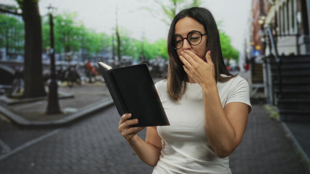 Woman in round glasses and white tshirt holding a black notebook and covering her mouth with her hand while yawning on a cobbled city street by a canal lined with bicycles; tiredness.