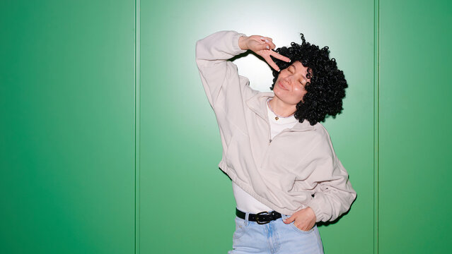 Young woman with curly wig and casual clothes making peace sign against a vibrant green wall