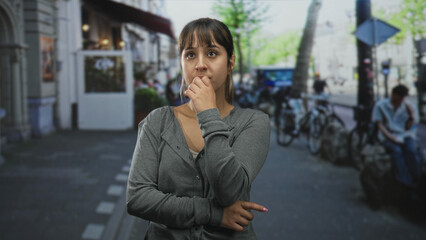 Fototapeta premium Woman with folded arms and visible hands standing on a city street near storefront and bicycles; pensive concern.
