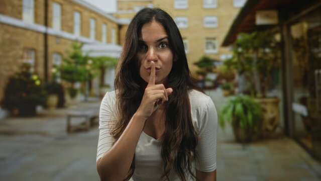 Woman finger to lips for silence in building courtyard with long hair and direct stare; silence restraint.