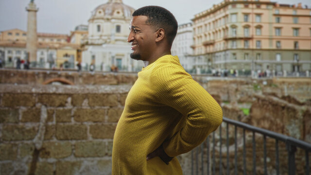 Young black man in yellow sweater holding hand on lower back for backache, wincing and bending slightly against a stone railing at a building; discomfort resilience.