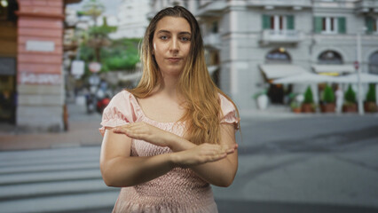 Fototapeta premium Woman crosses arms in an x over her chest with hands visible on a street crosswalk by sidewalk cafe umbrellas, wearing pink dress; defiance refusal.