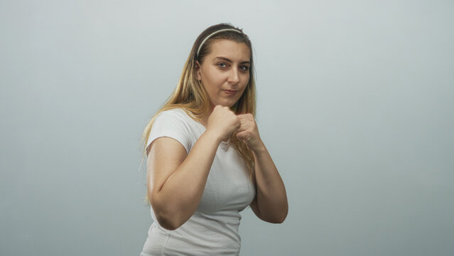 Woman with fists raised in punching motion, boxing guard stance, bare hands visible, wearing white tshirt and headband in studio; self defense confidence.
