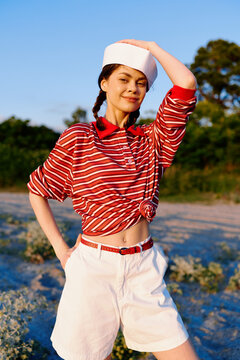 Young woman in a red striped shirt and white shorts poses on a sunny beach, smiling confidently while shading her eyes with a sailor hat. Casual lifestyle portrait, outdoor scene, summer fashion.
