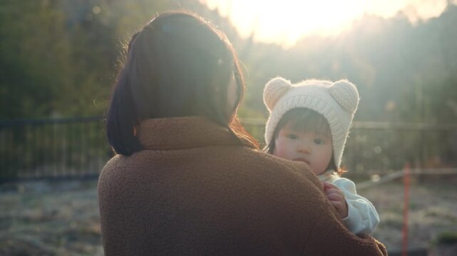 Back view of happy Asian mother holding her cute little daughter looking over shoulder at park, sweet baby girl wearing bear ear hat in mom arms during warm sunset for family travel vacation