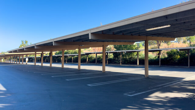 A long row of empty covered parking spaces under a large carport structure with support pillars next to green trees on a bright sunny day under a clear blue sky
