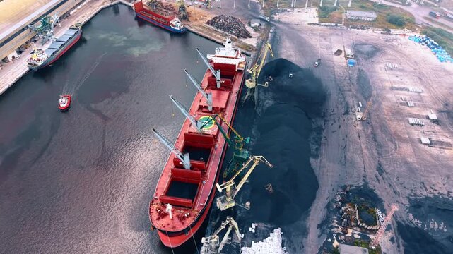 The large red vessel standing in the industrial bulk cargo terminal of the port. Geared bulk carrier with charcoal which is being transferred to the adjacent dockside piles.