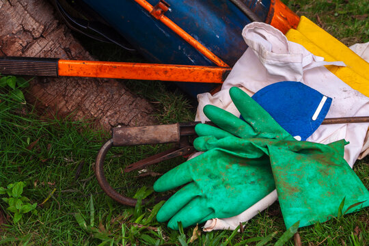 A pile of protective gardening and farming equipment lying on green grass near a tree trunk, including green chemical-resistant rubber gloves, a blue face mask, yellow cloth, 