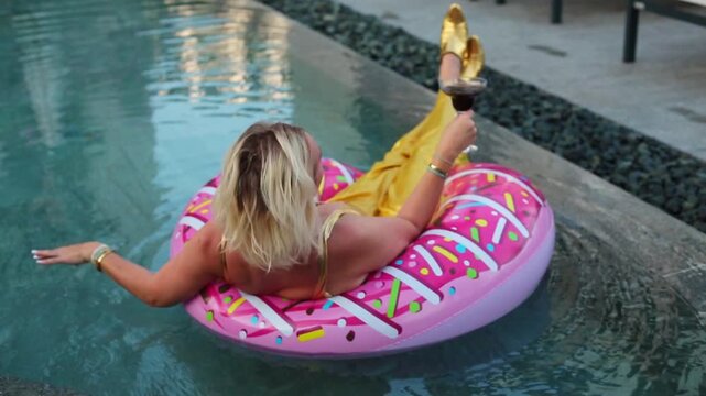 A blonde woman in a yellow swimsuit floats on a giant pink donut inflatable ring in a turquoise pool, relaxing with one hand raised to her face in a playful pose