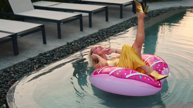 A blonde woman in a yellow swimsuit floats on a giant pink donut inflatable ring in a turquoise pool, relaxing with one hand raised to her face in a playful pose