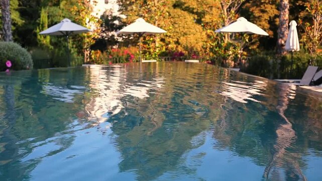 Water ripples and spills gently over the curved stone edge of a luxury infinity pool, with blurred sunloungers and lush colorful garden visible in the soft background