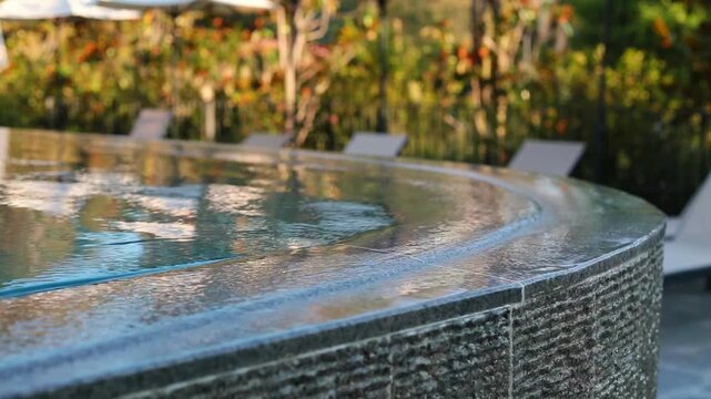 Water ripples and spills gently over the curved stone edge of a luxury infinity pool, with blurred sunloungers and lush colorful garden visible in the soft background