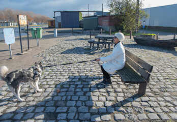 Woman in winter clothes sitting on a park bench, holding a leash for her husky dog standing on cobblestones next to her by a canal on a sunny day © Ekaterina