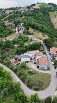 Vertical aerial drone view of rural houses with orange roofs by winding countryside road surrounded by green hills and trees in Italy region landscape.
