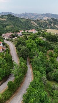 Vertical aerial drone view of rural houses with orange roofs by winding countryside road surrounded by green hills and trees in Italy region landscape.