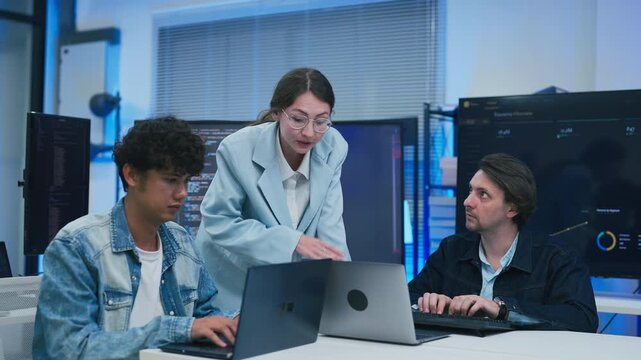 collaborative AI development team in modern tech office: young male developer coding on laptop while senior colleague leans over discussing code and data visualizations on multiple monitors	