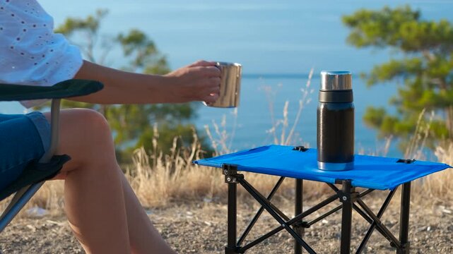 Drinking from a thermos at a campsite. Woman is reaching for a metal cup on a blue camping table with a black thermos on it. The sea and the coastline are in the background