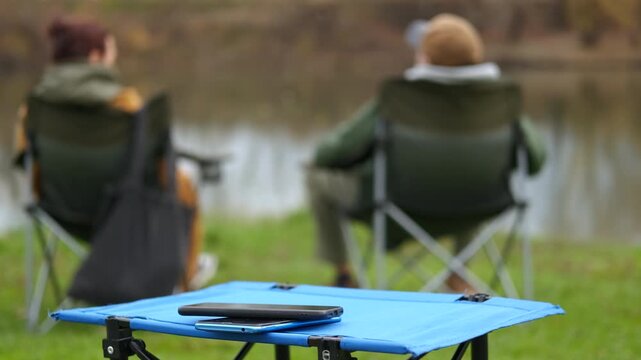 People fishing and relaxing near a lake. Two friends relaxing on folding chairs while fishing together at a tranquil lake, with their smartphones resting on a small portable table in the foreground