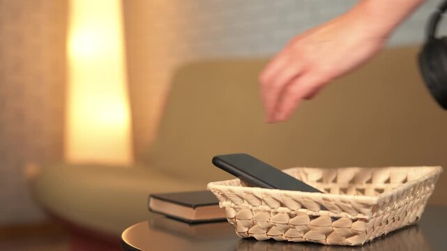Woman choosing book for digital detox at home. Relaxed woman putting her smartphone and headphones into a basket, reading a book instead for a evening at home, embracing the concept of digital detox