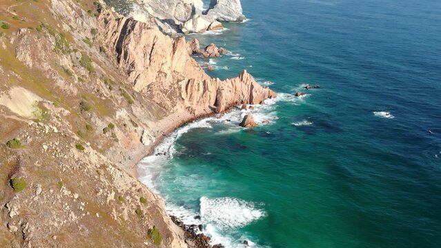 Aerial view of rugged coastal cliffs beside turquoise ocean waves and rocky shoreline in bright daylight . Media