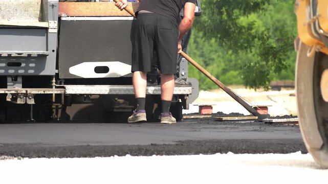 Worker spreads fresh asphalt on a road using a long-handled tool. He wears a black shirt and shorts. He concentrates intently on the task. Truck and road roller are nearby