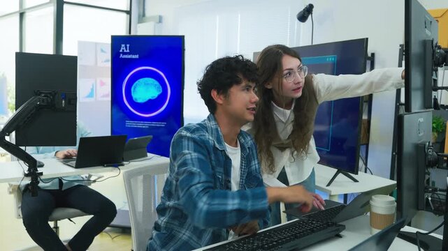collaborative AI development team in modern tech office: young male developer coding on laptop while senior colleague leans over discussing code and data visualizations on multiple monitors	