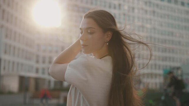 Young woman on urban rooftop shielding face with raised hand while long hair streams in wind under warm sunlight, wearing wide-brimmed hat and casual fashion near tall city buildings