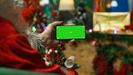 Fototapeta premium Santa Claus reads children's letters on a green screen smartphone in a workshop decorated with Christmas ornaments and a festive holiday atmosphere.
